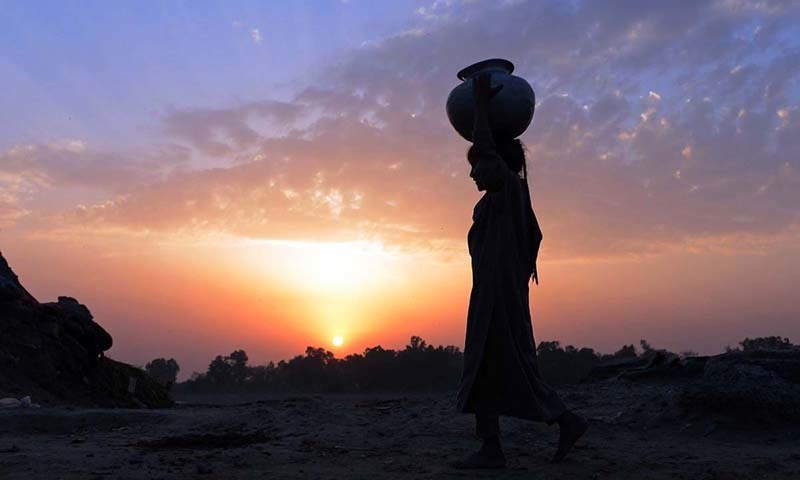 Description: A young girl carries a water pot over her head as she heads for home during sunset on the outskirts of Lahore, Punjab. — AFP/File
